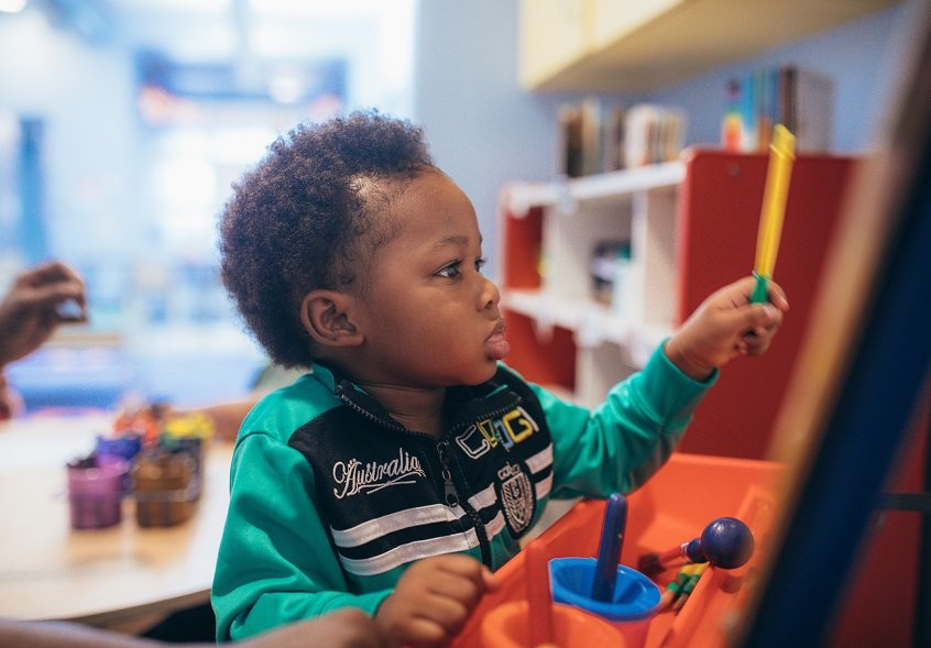 A young child in a green, black, and white jersey stands in front of an easel playing with paint brushes and art tools.