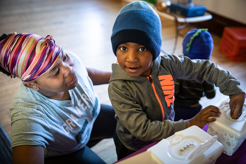 Image: A young child stands beside his caregiver, smiling happily together.