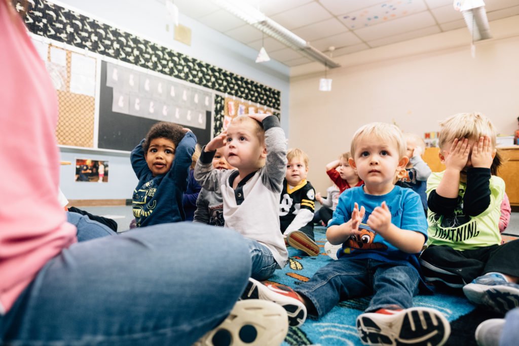 Picture: A group of young children sit in front of their teacher with their hands happily raised.