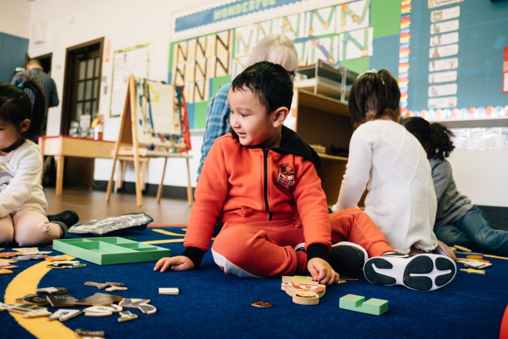 Picture: Young children sitting on a carpet, playing with small toys and cards. In the center, a young boy in orange looks off to the left.