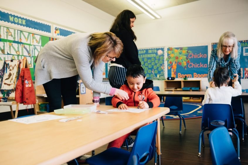 Image: A young child sits at a desk as an early learning professional bends over to help them with a project.