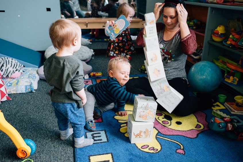 Image: An early childhood watches as three young children play with blocks and toys around her, as one child knocks down a giant block tower.