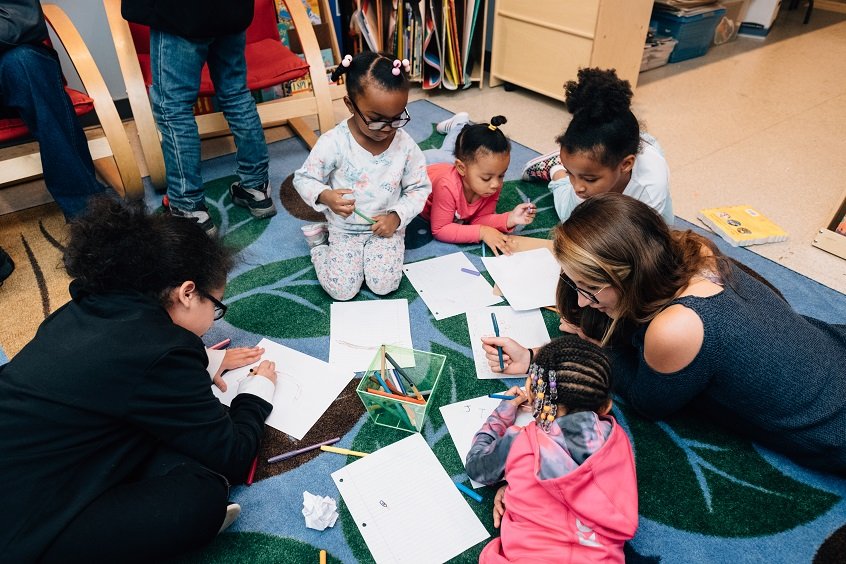 A group of young children sit together in a circle on the ground, writing on pieces of paper and talking to an early learning professional.