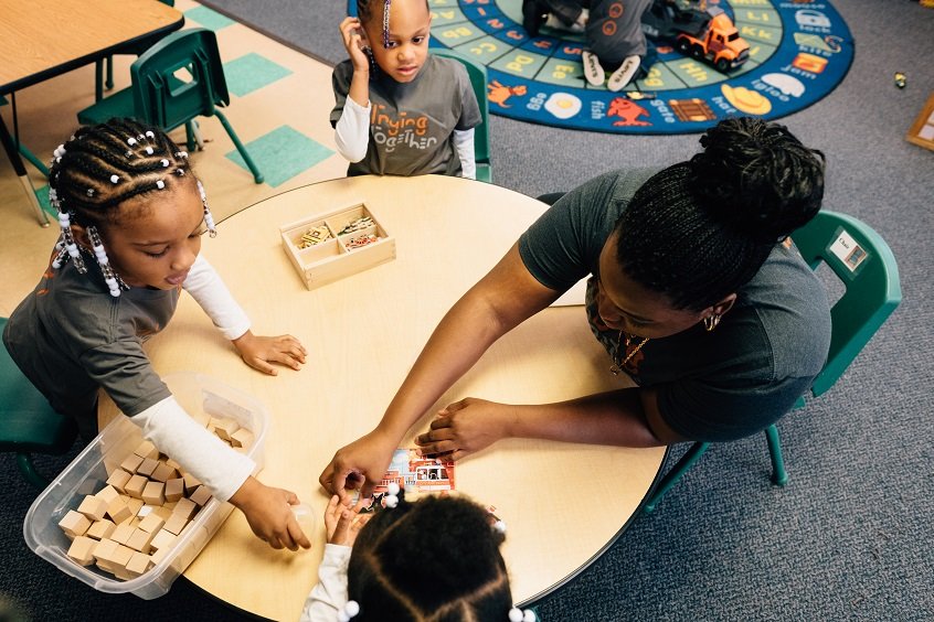 Image: An early learning professional sits on the ground with three children, playing together with small wooden toys.