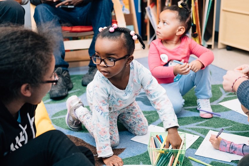 Image: Four young children sit together on the floor, all playing with different colored pencils, papers, and toys.