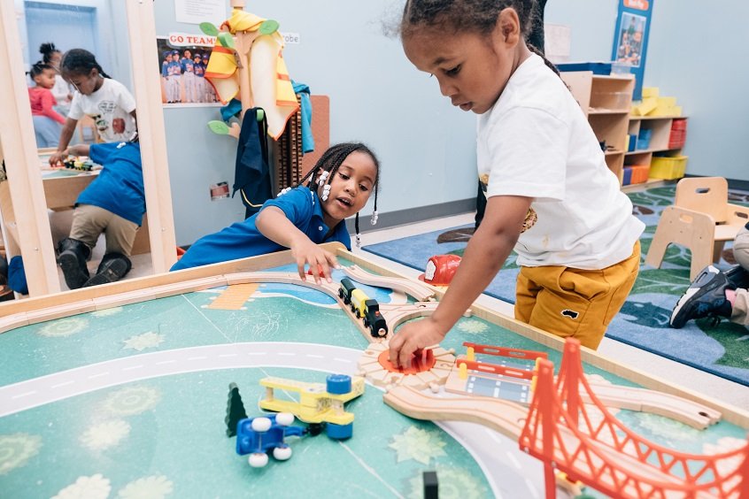 Image: Two young children play together at a colorful table, both holding small toys in their hands.