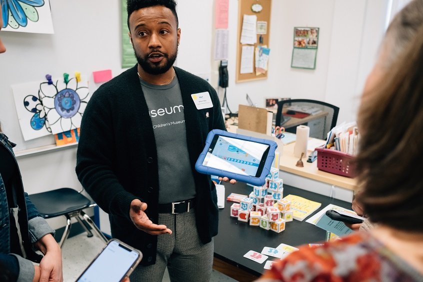 Image: A professional presents in to a small crowd, holding a tablet to demonstrate strategies for using tech.