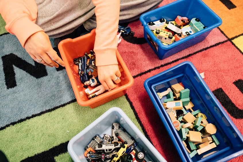 Image: An early learning professional sorts colorful toy pieces into four small boxes on the floor.