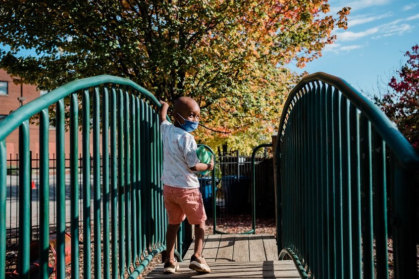 Image: A young child wearing a face mask looks back at the camera as he crosses a bridge.