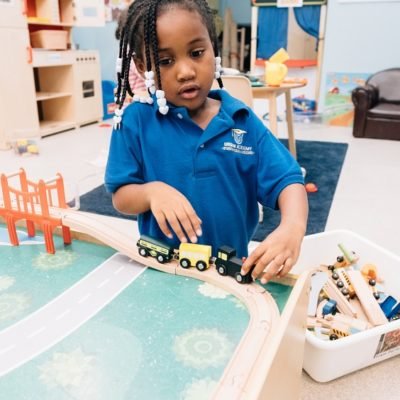 A young boy stands at a play table as he moves a toy train across a wooden track.