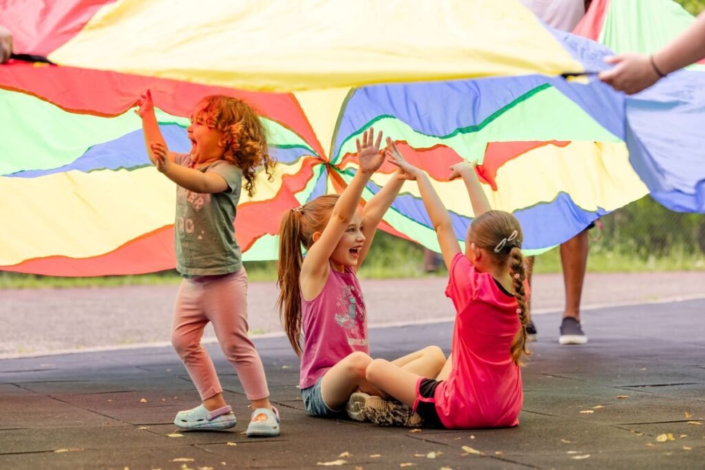 Three young girls sitting under the rainbow play tarp with huge smiles on their faces | Playing to Learn: Benefits of Play in Early Childhood