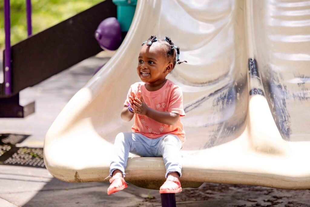 A young girl sitting on the edge of slide clapping and smiling | The Value of Play for Young Children